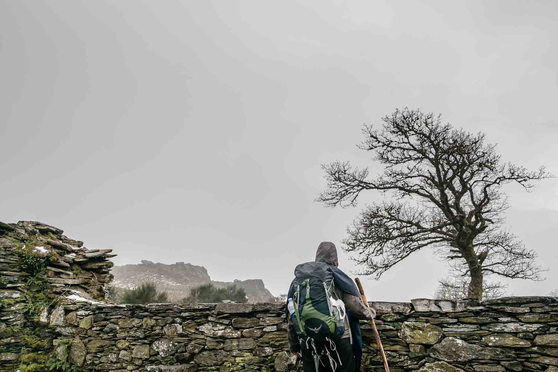 A man with a backpack standing next to a stone wall.